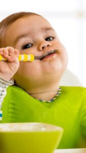 Um bebê com o rosto todo lambuzado com uma colher na boca